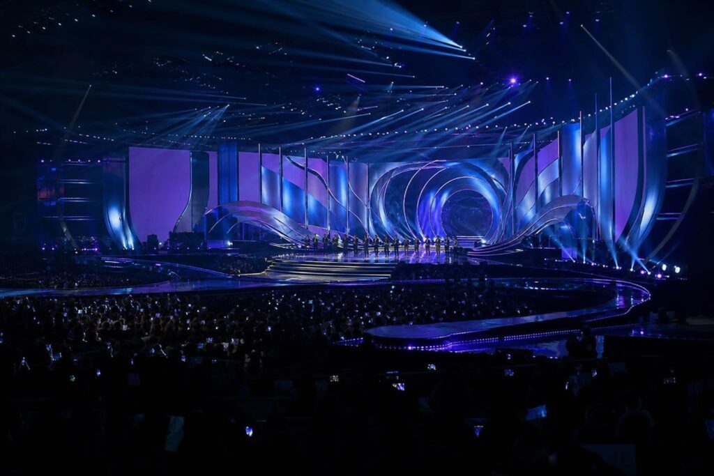 Panggung acara penghargaan musik K-pop dengan pencahayaan biru dan ungu serta penonton di Busan