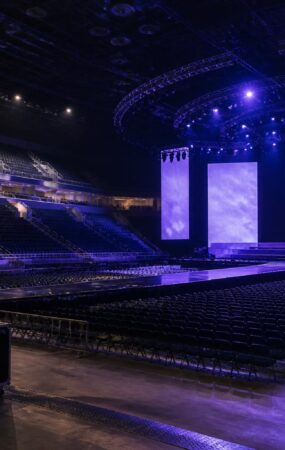Panggung konser arena yang kosong dengan pencahayaan biru dan ungu serta perlengkapan produksi di sisi belakang