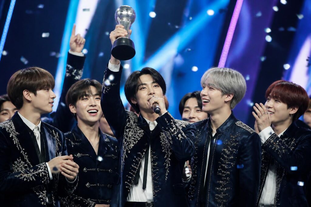 Grup K-pop merayakan kemenangan lagu terbaru mereka di atas panggung acara musik dengan trofi dan konfeti.