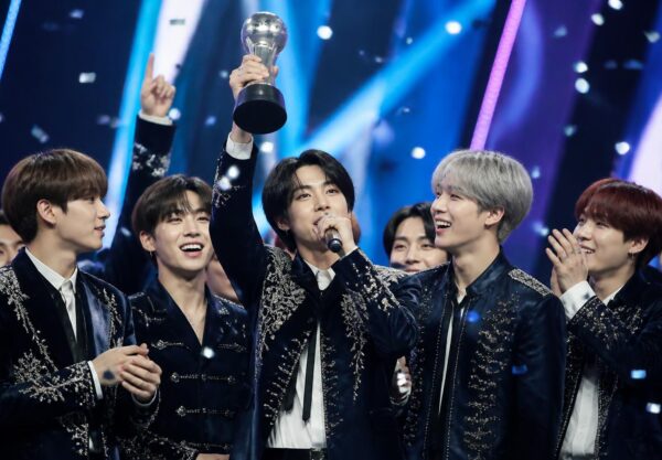 Grup K-pop merayakan kemenangan lagu terbaru mereka di atas panggung acara musik dengan trofi dan konfeti.