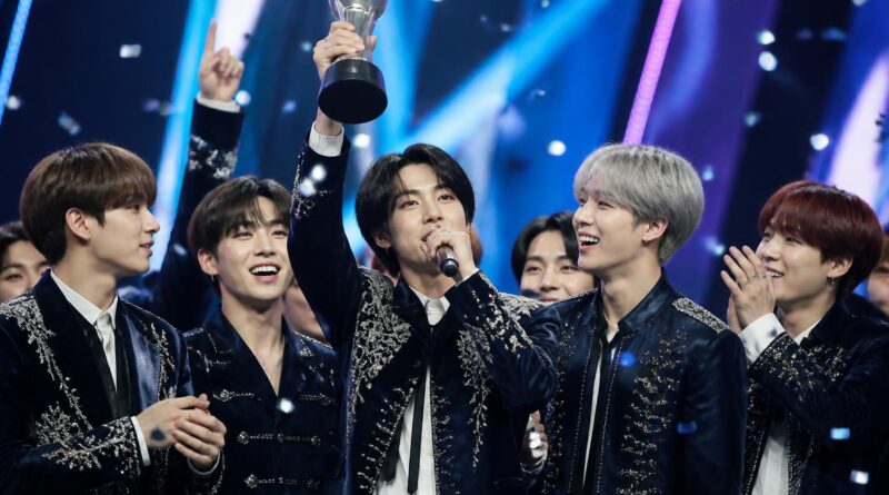 Grup K-pop merayakan kemenangan lagu terbaru mereka di atas panggung acara musik dengan trofi dan konfeti.