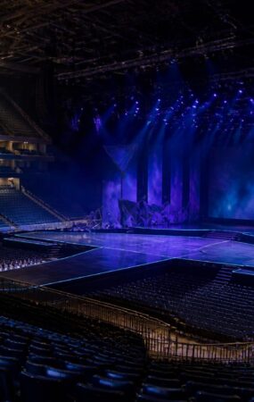 Panggung konser di arena dalam ruangan dengan pencahayaan biru dan ungu serta deretan kursi penonton yang kosong