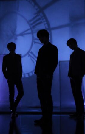 Siluet sekelompok performer dalam suasana studio bernuansa biru dan ungu yang menggambarkan hitung mundur comeback musik